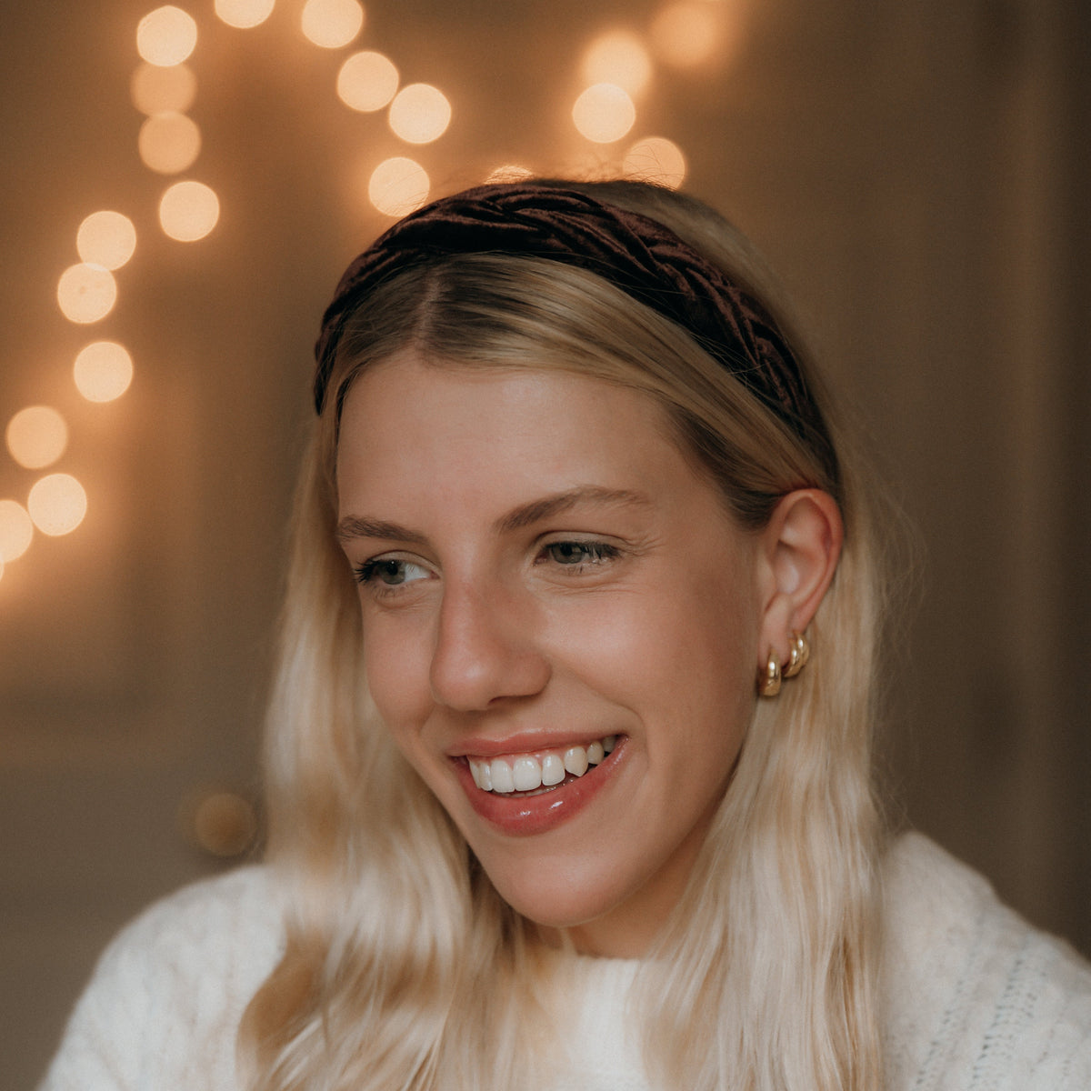 Woman with long blonde hair wearing a headband, smiling in front of warm string lights.