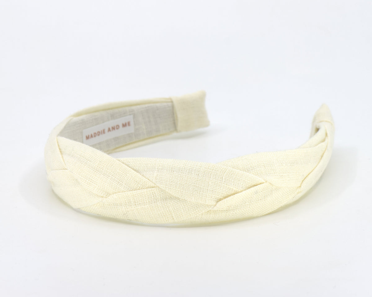 A pale cream woven fabric headband with a braided crisscross pattern, featuring a visible 'MADDIE AND ME' brand label, set against a plain white background.