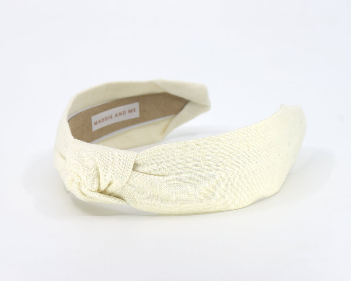 A cream-colored fabric headband with a twisted knot detail, displayed on a clean white background. The inner label shows the brand 'MADDIE AND ME'.