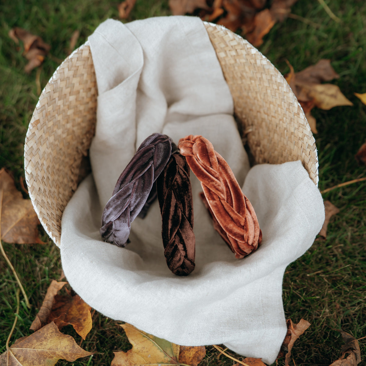Woven basket with velvet headbands on a white cloth and dried leaves on grass.