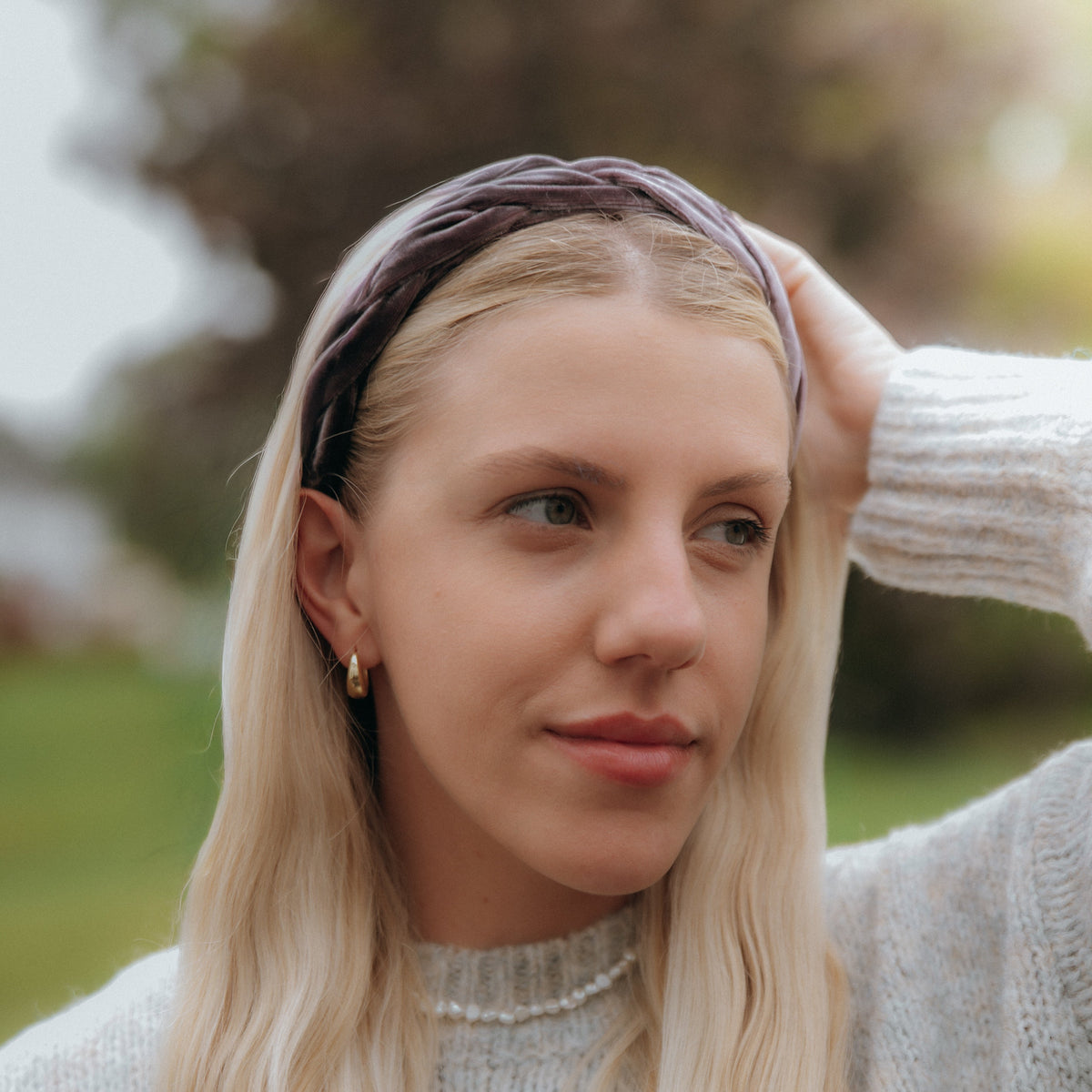 Woman with long blonde hair wearing a velvet braided headband, standing outdoors with blurred trees in the background.