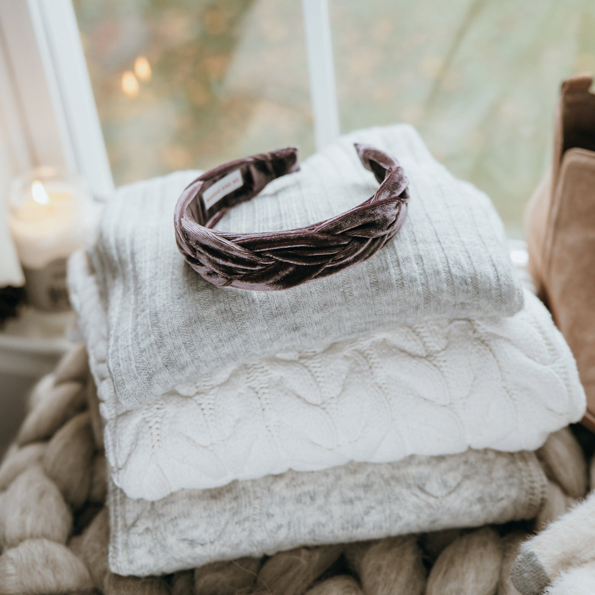 Velvet braided headband on a stack of sweaters on a decorative basket on a textured surface near a window.