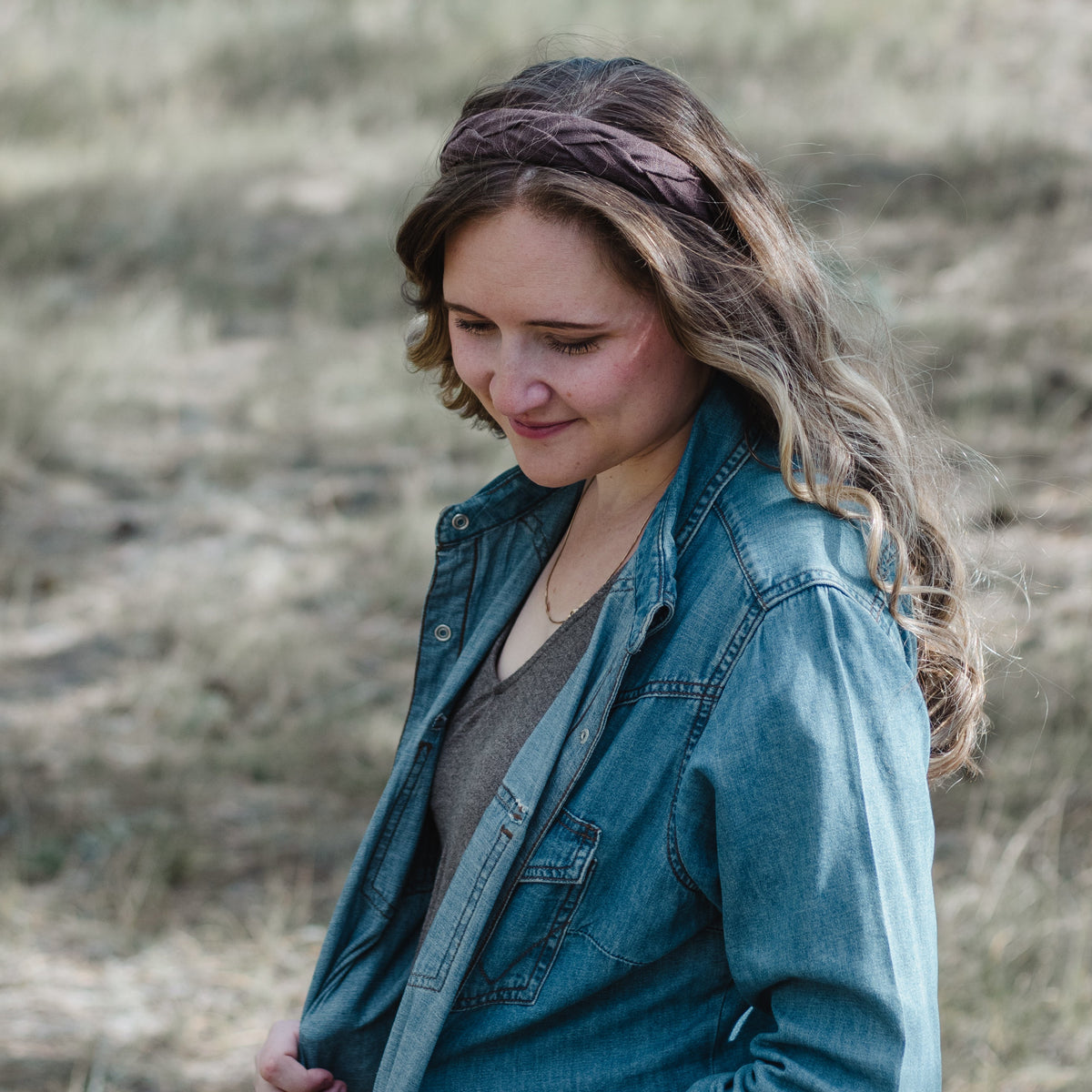 A woman wearing a blue denim button-up shirt over a brown top, standing outdoors in a dry grassy field. She has a brown braided headband on.