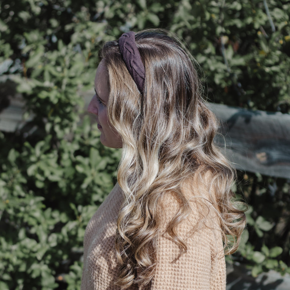 A woman in profile wearing a chocolate brown fabric headband and a beige waffle-knit sweater, set against a blurred background of green foliage.