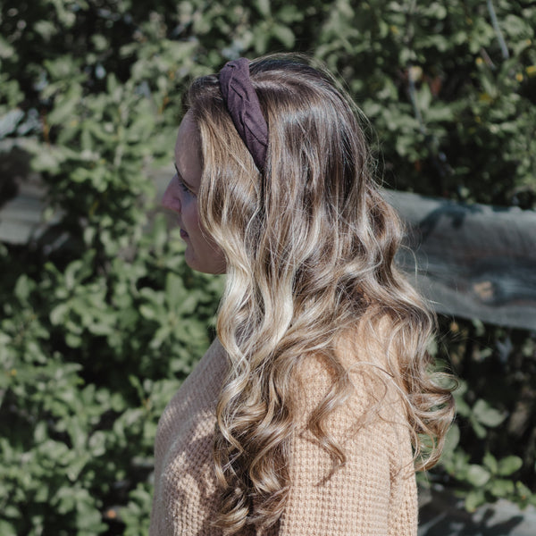 A woman in profile wearing a chocolate brown fabric headband and a beige waffle-knit sweater, set against a blurred background of green foliage.