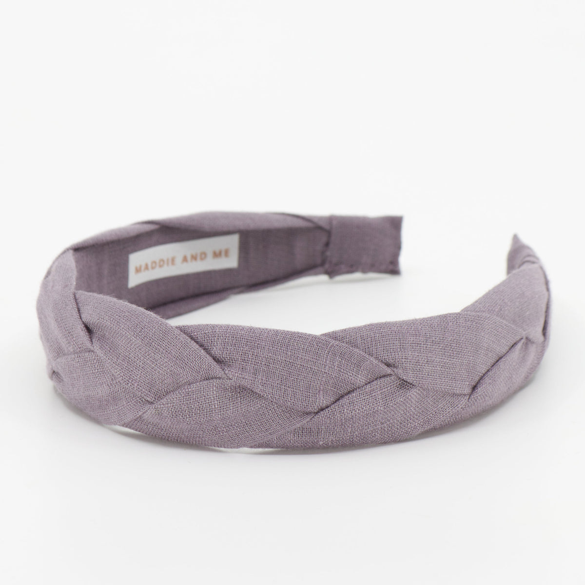 A wide braided fabric headband in a muted dusty purple color, featuring a small interior label that reads 'MADDIE AND ME'. The headband is photographed on a seamless white background.