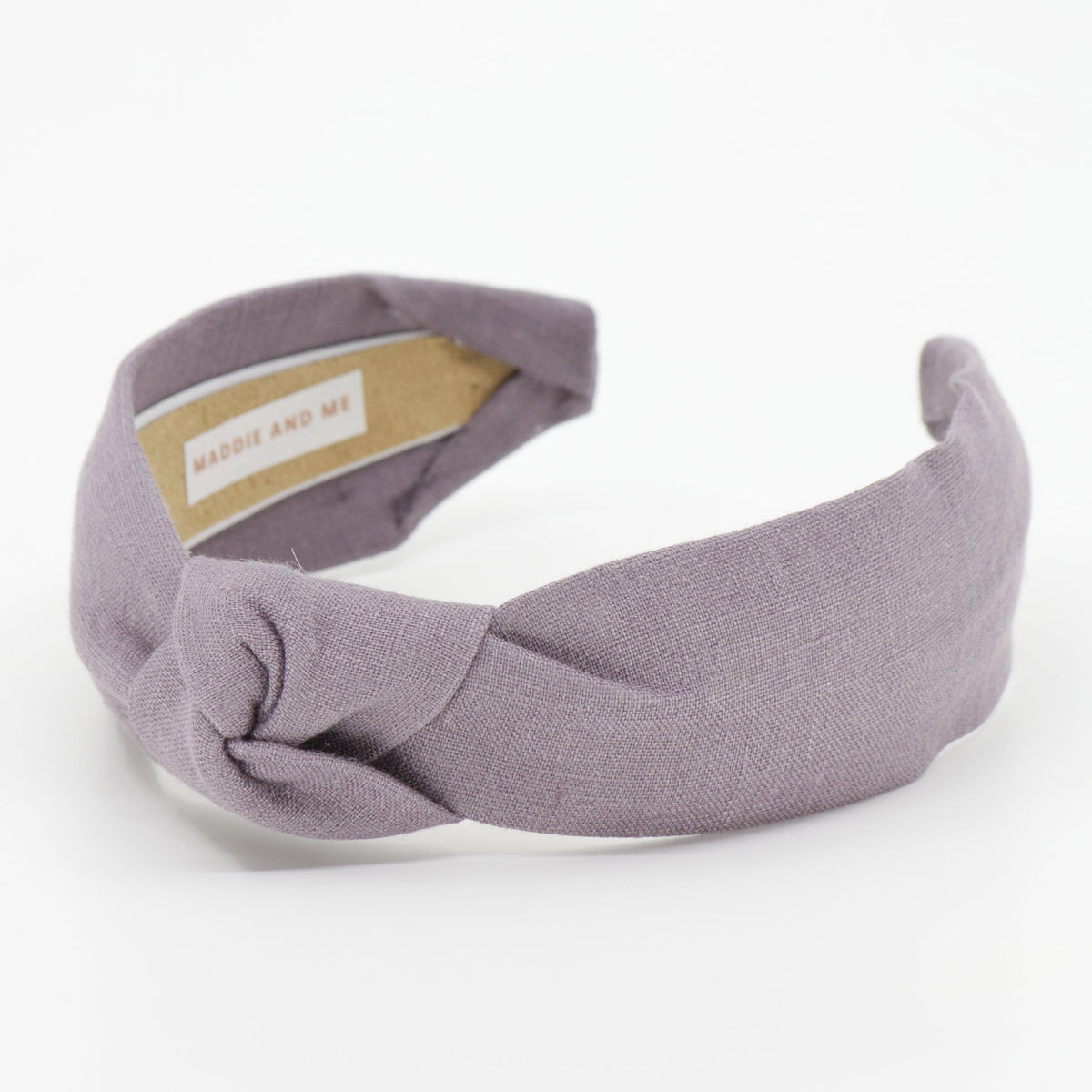 A dusty purple twisted fabric headband with a knotted center, shown on a clean white background. A small beige paper label is visible inside the headband.