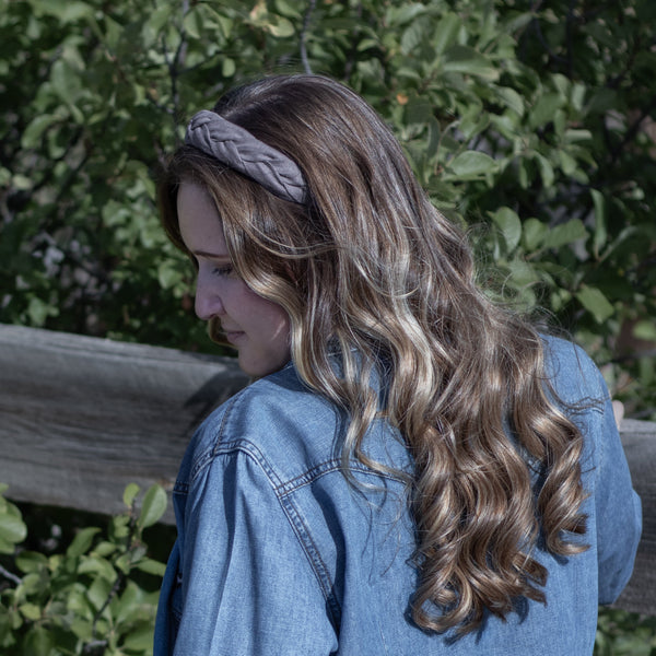A woman with long, wavy brown hair wearing a mauve braided headband and a blue denim shirt, photographed outdoors against a background of green foliage and a wooden fence.