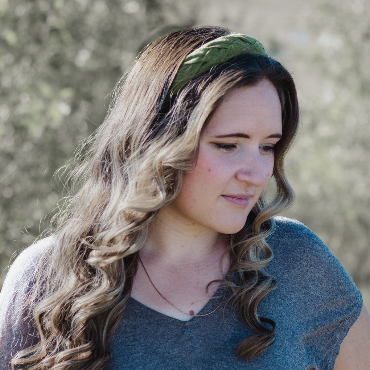 A woman with long wavy brown hair wearing an olive green fabric headband and a heather gray t-shirt, photographed outdoors with a blurred natural background.