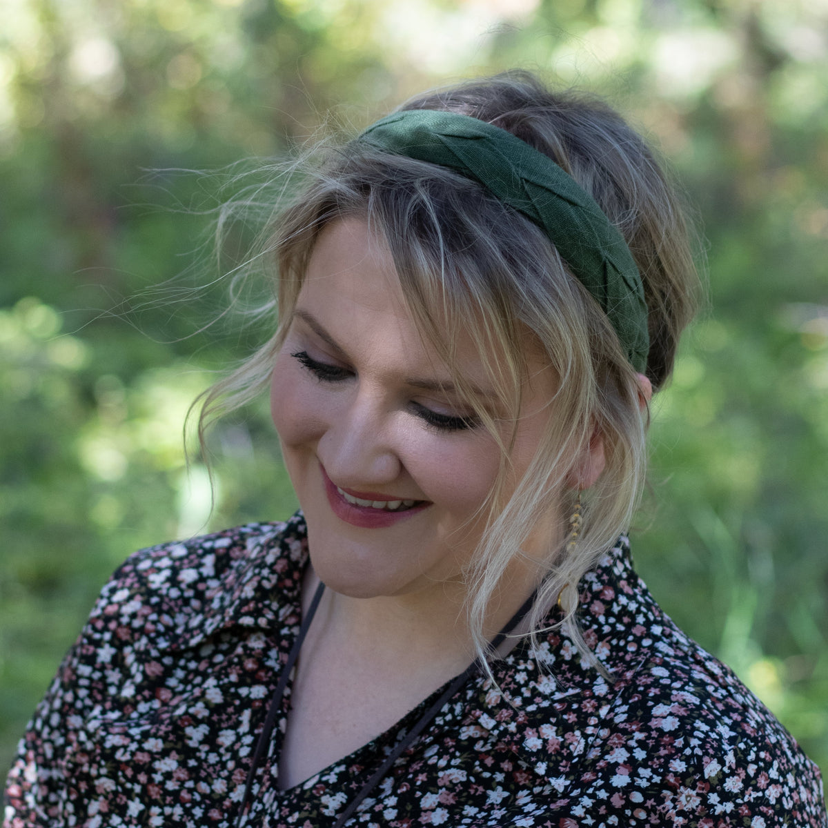 A woman outdoors wearing a dark green wide fabric headband and a black floral blouse, looking down with a soft smile. The background is a blurred green natural setting.